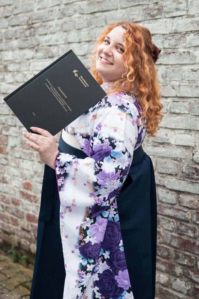 Frau mit rotem Haar in einem Kimono und Hakama hält ein Abschlusszeugnis und lächelt.