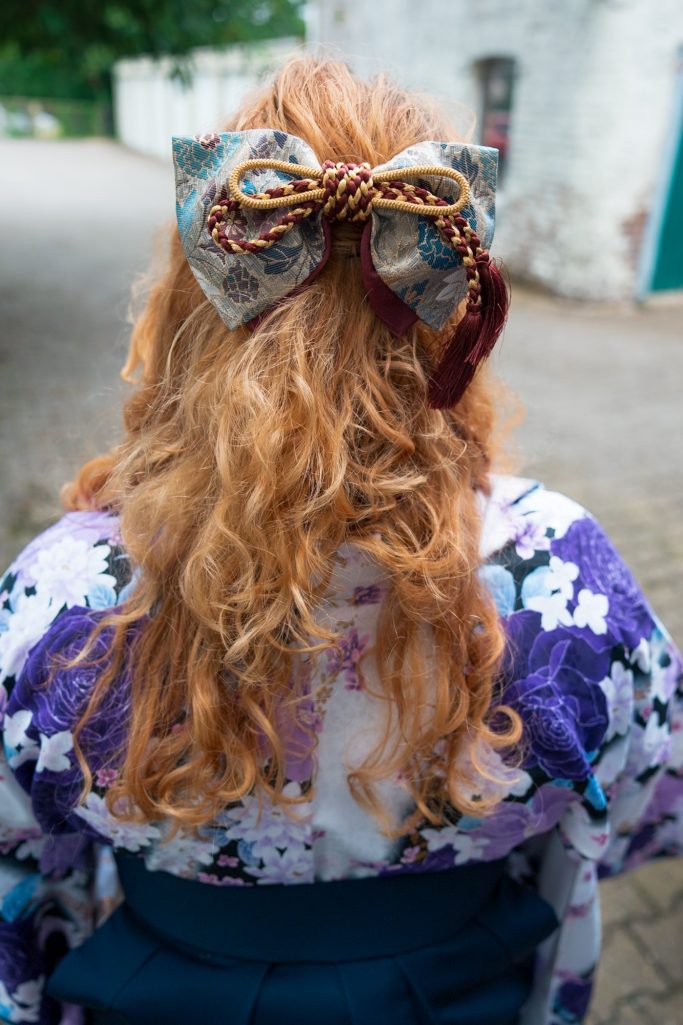 Frau mit lockigem, rotblondem Haar und großer, farbiger Schleife im Rücken trägt ein Kimono.