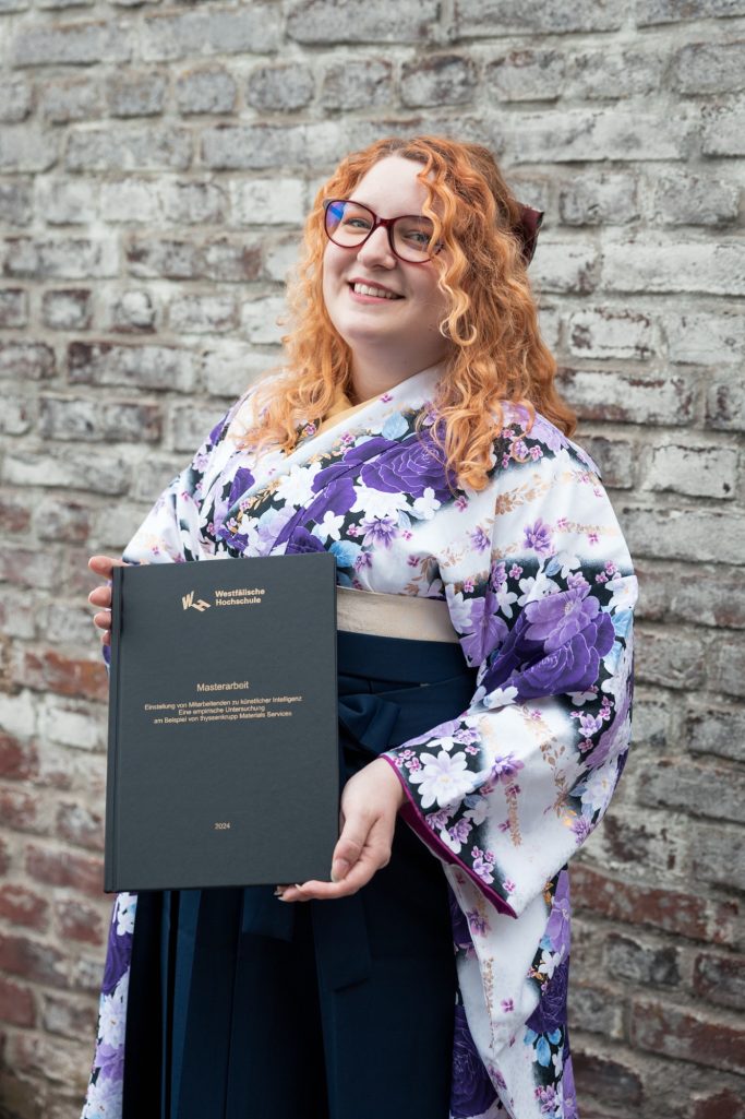 Eine lächelnde Frau in einem Kimono und Hakama hält ihre Masterarbeit vor einer Steinmauer.
