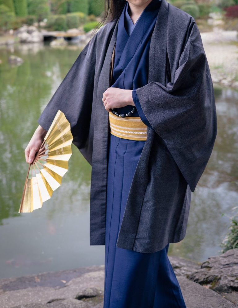 Ausgeh-Männerkimono Front Person in traditioneller Kleidung hält einen goldenen Fächer vor einem Teich.