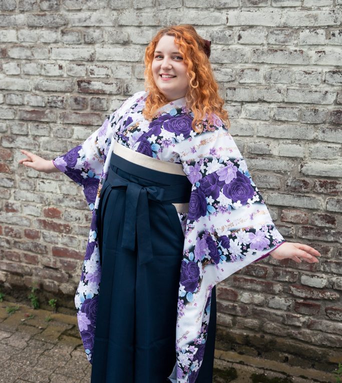 Eine lächelnde Person in einem lila Furisode mit Hakama vor einer Steinmauer.